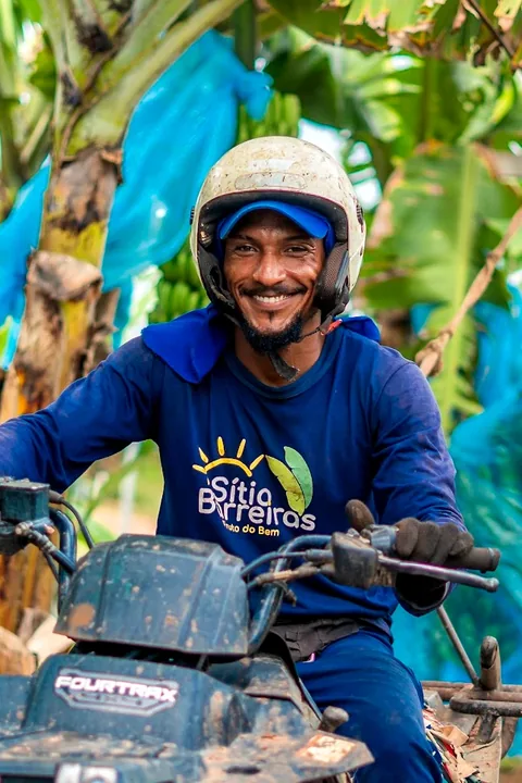 Colaborador sorrindo, de capacete, no quadriciclo entre as bananeiras